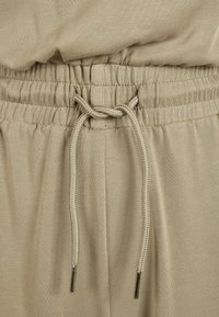 Pantalons de survêtement beige avec une taille élastique, dotés d'un cordon de serrage avec des embouts métalliques et d'une texture froncée autour de la taille.