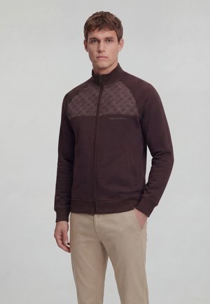 Młody mężczyzna ubrany w ciemnobrązową kurtkę zapinaną na zamek z wzorzystą górną częścią na klatce piersiowej oraz jasnobeżowe spodnie, stojący na tle jednolitego szarego tła.