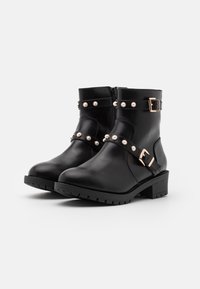 Botas de tobillo de cuero negro con un tacón grueso, que presentan acentos de hebilla en tono dorado y tachuelas decorativas similares a perlas en las correas.