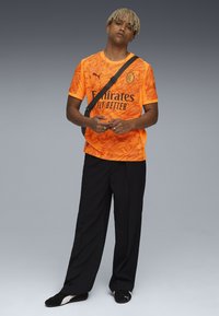 Maglietta sportiva arancione brillante con un motivo testurizzato, logo nero stampato, abbinata a pantaloni scuri a vestibilità larga e scarpe nere.