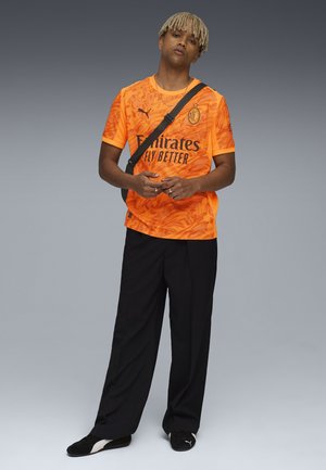 Kirnu oranssi urheilupaita, jossa on tekstuurikuvio, musta painettu logo, yhdistettyinä tummiin löysiin housuihin ja mustiin kenkiin.