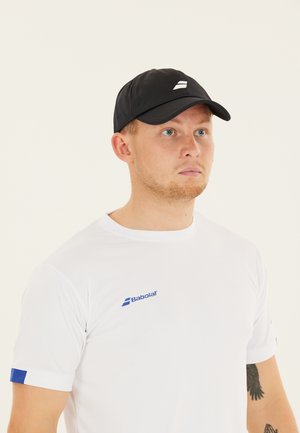 Ung mand iført en sort kasket og en hvid Babolat sportst-shirt, der kigger let mod højre, med en delvis tatovering synlig på hans arm.