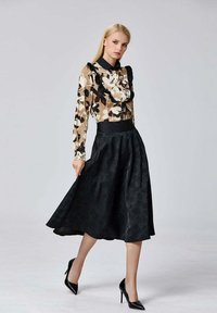 La blouse présente un motif floral dans des tons terreux, des détails en volants et un col noir ; elle est associée à une jupe noire mi-longue et des escarpins noirs.