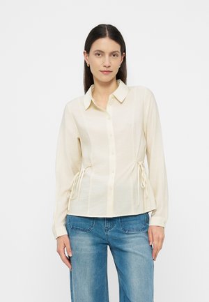 Tumevate juustega naine, kes kannab kreemjat pika käisega nööbitavat pluusi küljetilgadega ja siniseid teksapükse, seisab tavalise tausta ees.