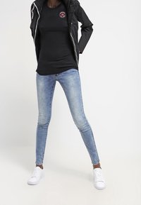 Mustat vetoketjullinen takki logolla, musta toppi, vaaleansiniset skinny-farkut ja valkoiset lenkkitossut. Asusteessa on rento, istuva muotoilu.