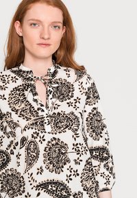 Top em padrão preto e branco com decote amarrado e mangas ¾, apresentando desenhos florais e paisley sobre um fundo claro.