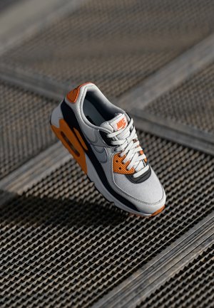 Białe, czarne i pomarańczowe buty sportowe Nike unoszące się nad metalową kratką, podkreślające szczegóły boku i górnej części.