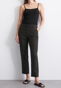 Débardeur noir ajusté, associé à un pantalon taillé vert foncé avec des poches latérales. Le modèle porte des sandales à glissière noires. Tissu lisse, design à jambes droites.