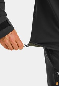 Black softshell jacket with a hand pulling a drawstring. Features include a zipper at the hem and contrasting green detail near the bottom.