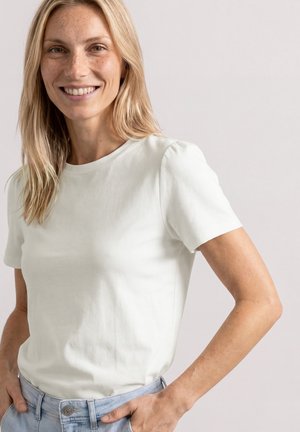 Uśmiechnięta kobieta z długimi blond włosami, ubrana w prostą białą koszulkę z krótkim rękawem i jasnoniebieskie dżinsy, stojąca na jasnym tle.