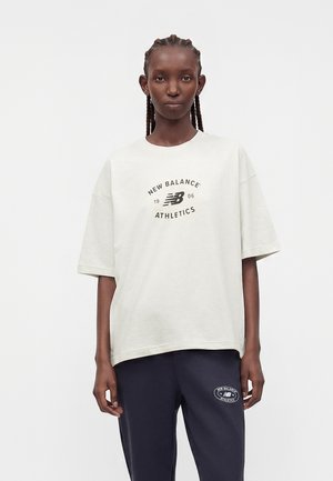 Frau trägt weißes New Balance Athletics T-Shirt und dunkelblaue Jogginghose mit passendem Logo, steht vor einfachem hellem Hintergrund.