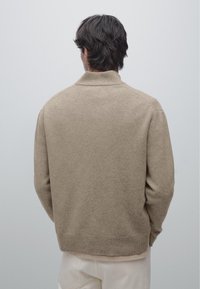Beige strikket sweater med høj krave, ribstrikkede manchetter og kant. Blød tekstur med en afslappet pasform, vist bagfra mod en neutral baggrund.