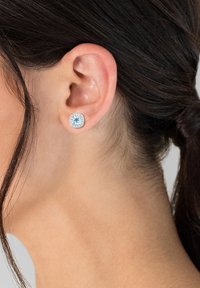 Runde silberne Ohrringe mit einem hellblauen zentralen Stein, umgeben von einem funkelnden Halo aus kleineren klaren Steinen. Glatte metallische Oberfläche.