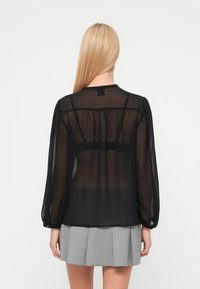 Zwarte doorschijnende blouse met lange mouwen, geplooide achterkant en een ronde halslijn. Gecombineerd met een lichtgrijze geplooide rok.