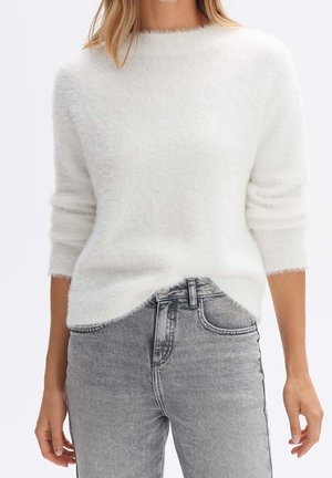 Femme portant un pull doux blanc et duveteux à manches longues associé à un jean en denim gris clair taille haute sur un fond uni.