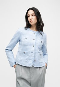 Γυναίκα με μακριά σκούρα μαλλιά φοράει μπλε ανοιχτό τζάκετ με υφή και ασημένια κουμπιά και ανοιχτόχρωμο γκρι παντελόνι με ρίγες, στέκεται μπροστά σε λευκό φόντο.