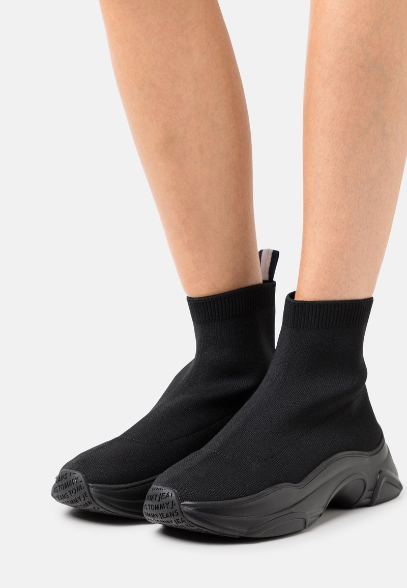 Baskets noires style chaussette avec semelles épaisses et texte « Tommy Jeans » sur les bouts, portées sur des jambes nues sur fond blanc.