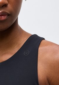 Κοντινό πλάνο στον ώμο και τον λαιμό ενός ατόμου που φοράει μαύρο τοπ χωρίς μανίκια με ένα μικρό κυκλικό λογότυπο κοντά στην κλείδα.