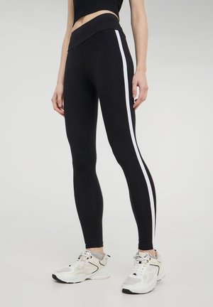Leggings neri con una fascia larga in vita, caratterizzati da una striscia bianca laterale. Tessuto morbido e elastico e design aderente. Indossati con sneaker bianche.