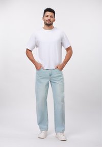 Camiseta blanca de manga corta combinada con unos vaqueros holgados de color azul claro. El modelo está de pie con las manos en los bolsillos, llevando zapatillas blancas.