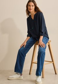 Marineblouse met V-hals en trekkoord, gecombineerd met blauwe denim jeans. Witte sneakers met kastanjebruine accenten. Achtergrond met een houten kruk.