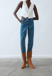 Gilet bianco corto con scollo a V profondo e bottoni, abbinato a jeans blu a vita alta e stivali con tacco alti color carne. Tessuti e finiture morbide.