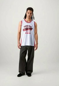 Noor noormees, kellel on dredsid, kannab valge-punast ilma varrukateta Santa Cruzi korvpallisärki ja laiu musti pükse, seismas ühtlase tausta ees.