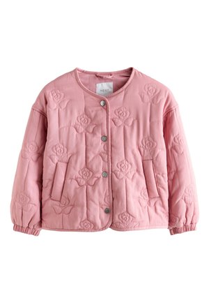 Ung pige står indendørs med hænderne i lommerne på vidbukser, iført en lyserød quiltet jakke og hvide sneakers på trægulv.