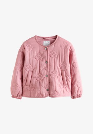 Ung pige står indendørs med hænderne i lommerne på vidbukser, iført en lyserød quiltet jakke og hvide sneakers på trægulv.