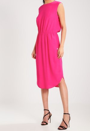 Vestido sin mangas de color rosa brillante hasta la rodilla con cintura ceñida, combinado con sandalias negras de tacón alto y tiras, en una persona de pie.
