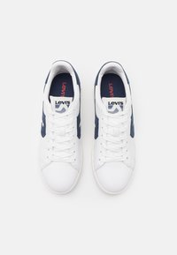 Białe sneakersy z granatowymi akcentami, charakteryzujące się gładką skórzaną teksturą, sznurowanym zapięciem oraz markową wkładką z logo na boku.