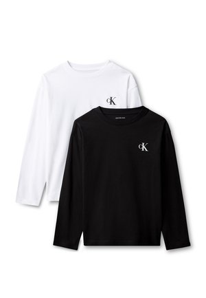 Dreng med mørkebrunt hår iført sort langærmet skjorte, hvid sweatshirt bundet om livet, mørke bukser og hvide sneakers, står foran en grå baggrund.