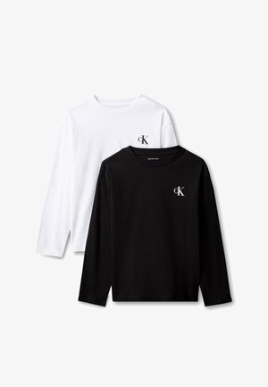 Jongen met donkerbruin haar draagt een zwart shirt met lange mouwen, een witte trui om de taille geknoopt, donkere broek en witte sneakers, staand tegen een grijze achtergrond.