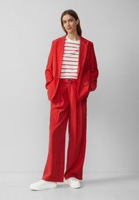 Roter maßgeschneiderter Blazer mit passenden lockeren Hosen, kombiniert mit einem weißen Hemd mit roten Streifen. Dazu weiße Sneakers.