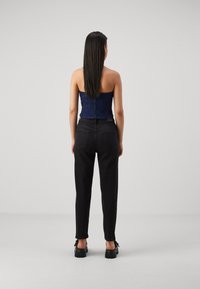 Kvinna med långt mörkt hår som bär en strapless marinblå topp, svarta högmidjade jeans och svarta klumpiga skor, stående mot en ljus enfärgad bakgrund.