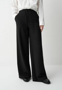 Een persoon in een witte, geplooide blouse en wijde zwarte broek staat tegen een neutrale achtergrond, met de handen in de zakken, en toont de outfit.