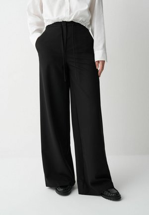 Een persoon in een witte, geplooide blouse en wijde zwarte broek staat tegen een neutrale achtergrond, met de handen in de zakken, en toont de outfit.
