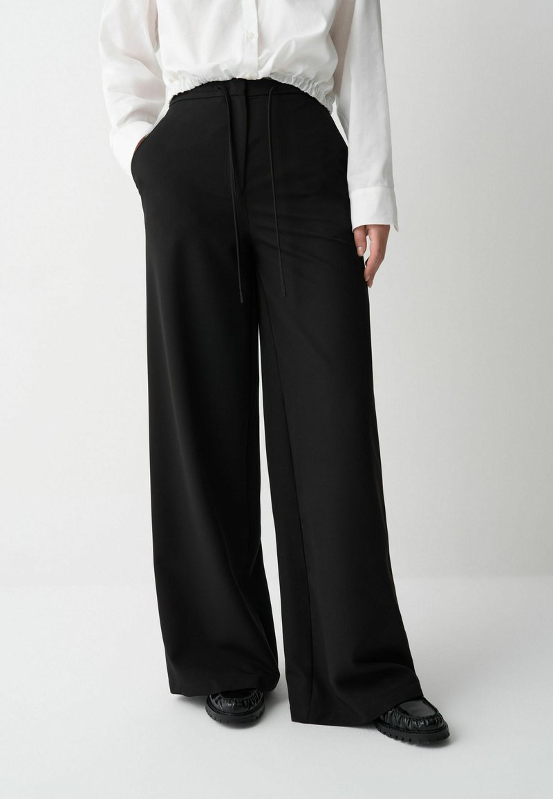 Een persoon in een witte, geplooide blouse en wijde zwarte broek staat tegen een neutrale achtergrond, met de handen in de zakken, en toont de outfit.