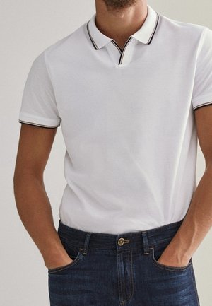 Homme portant un polo blanc à manches courtes avec un bord noir et un jean bleu foncé, les mains dans les poches, debout devant un fond uni.