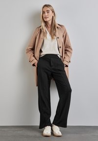 Trench-coat beige clair avec poches latérales, porté sur un pull crème et un pantalon noir ajusté. Des baskets blanches avec des accents bruns complètent le look.