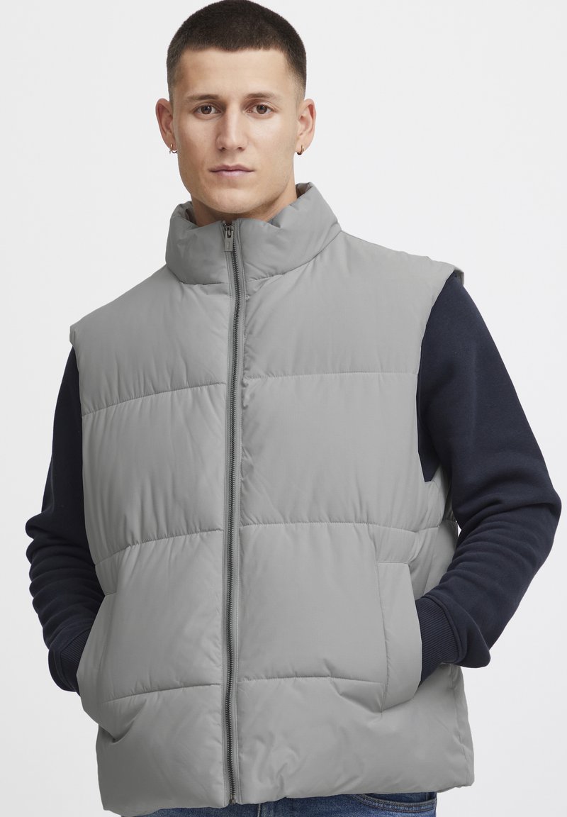 Graue gepolsterte Weste mit hohem Kragen, ausgestattet mit einem durchgehenden Reißverschluss und seitlichen Taschen. Über einem marineblauen Langarmshirt getragen, mit glatter Textur.