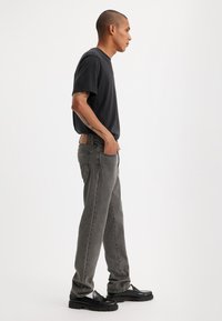 Grå denim jeans med avslappet passform, kombinert med en svart kortermet skjorte. Modellen står sidelengs, iført svarte platå-sko.