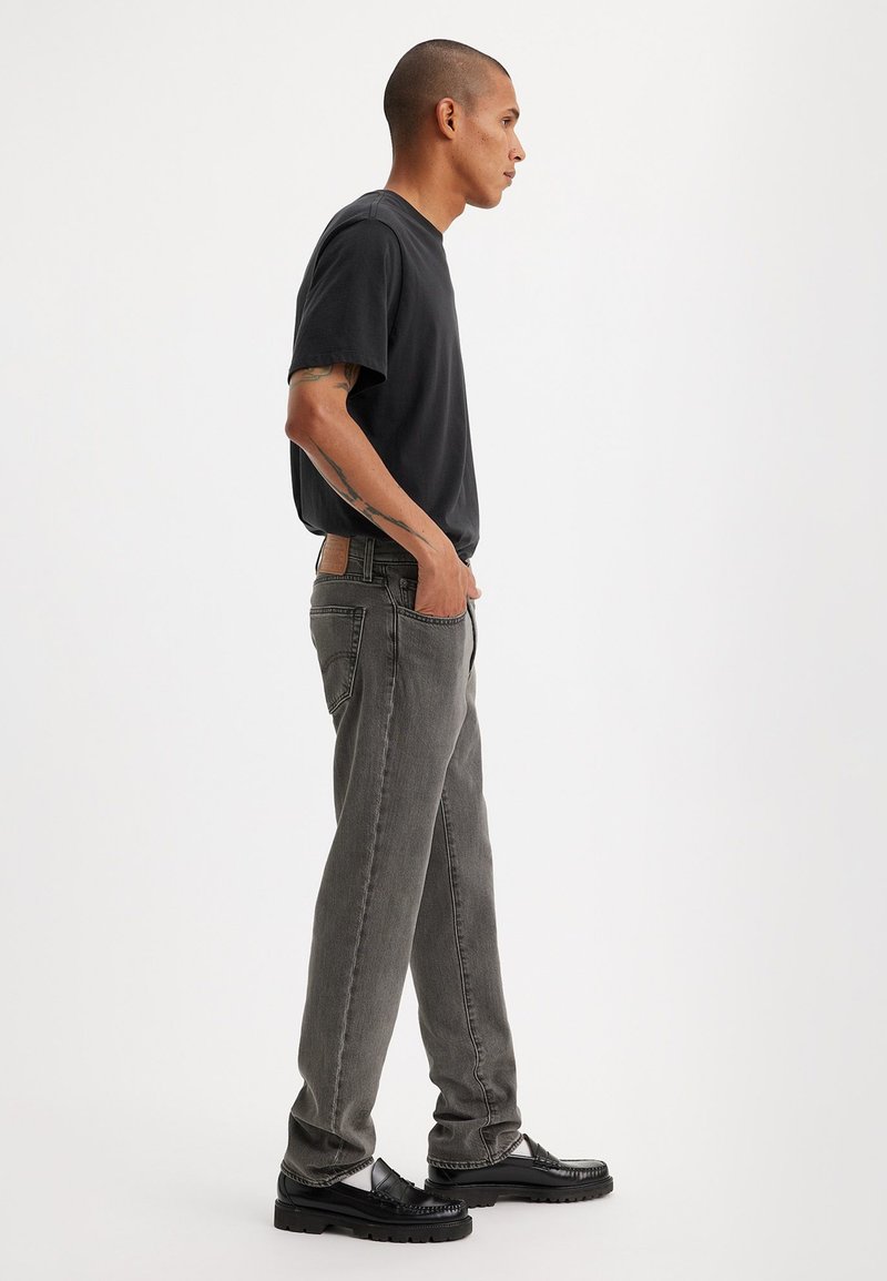 Jean en denim gris avec une coupe décontractée, associé à une chemise noire à manches courtes. Le modèle se tient de côté, portant des chaussures à plateforme noires.