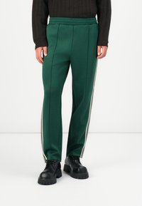 Pantaloni atletici verzi cu talie ajustată, cu două dungi albe pe laterale și pliuri. Purtați cu pantofi negri masivi pentru contrast.