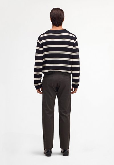 Homme de dos portant une chemise à manches longues rayée noir et blanc, un pantalon foncé et des chaussures noires devant un fond blanc.