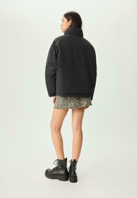 Donna che indossa una giacca a vento nera, una mini gonna leopardata e stivali platform neri con lacci, in piedi contro uno sfondo chiaro e semplice.