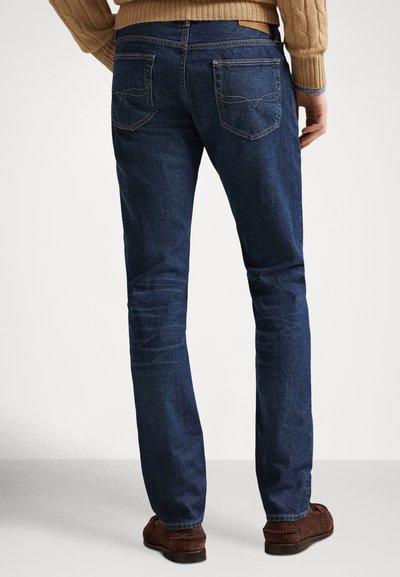 Mężczyzna ubrany w ciemnoniebieskie jeansy o prostych nogawkach, brązowe mokasyny z zamszu oraz beżowy sweter, stojący na jasnym, jednolitym tle.