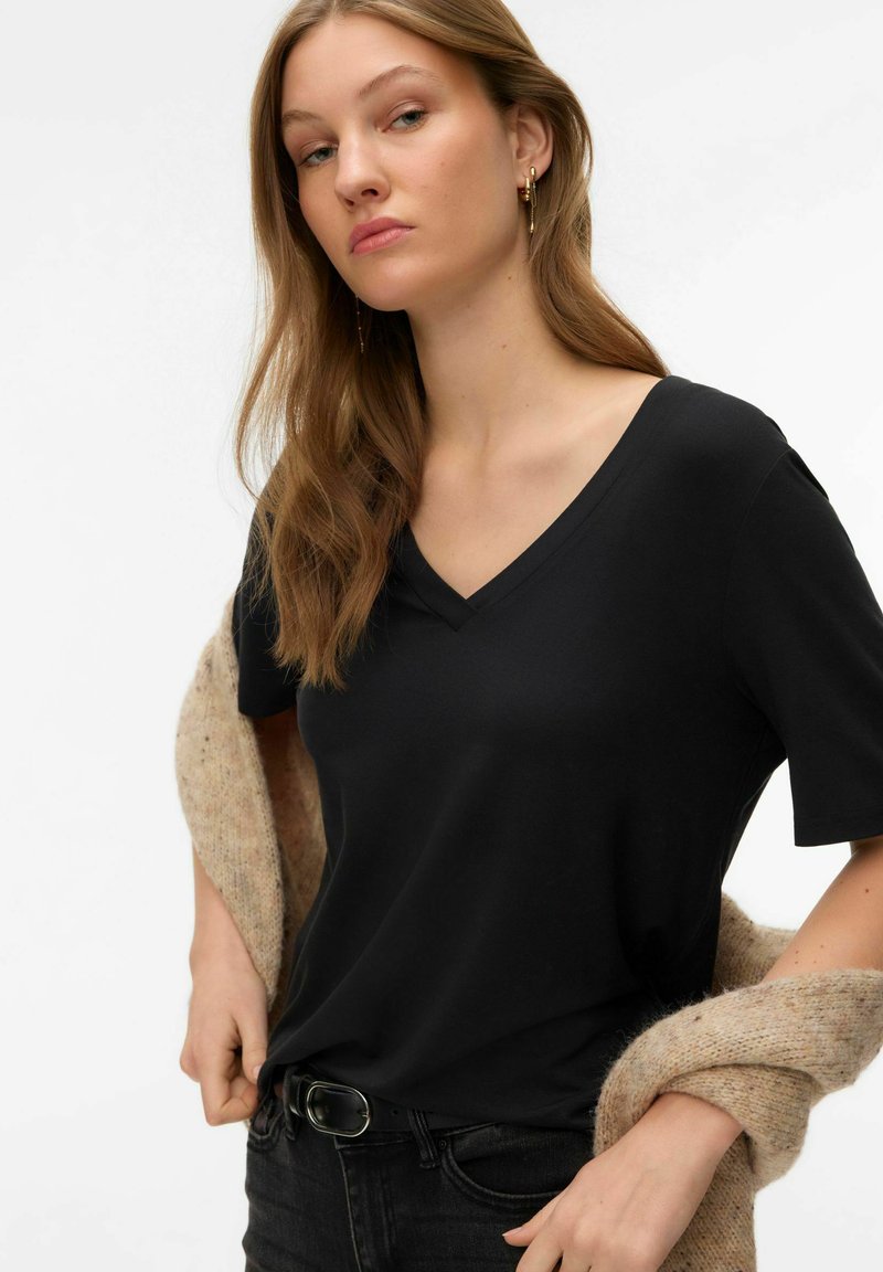 Camiseta negra de cuello en V en tela suave, manga corta, combinada con un suéter beige de punto sobre el brazo, usada con jeans oscuros.