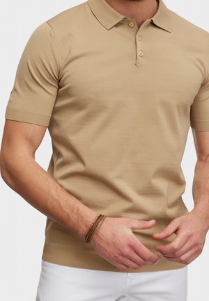 Polo beige a maniche corte indossata da un uomo, con tre bottoni al collo, abbinata a pantaloni bianchi e braccialetti marroni.
