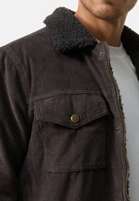 Zwarte corduroy jas met een gestructureerde, fleece gevoerde kraag. Voorzien van een ritssluiting aan de voorkant en een borstzak met een ronde gouden knoopdetail.